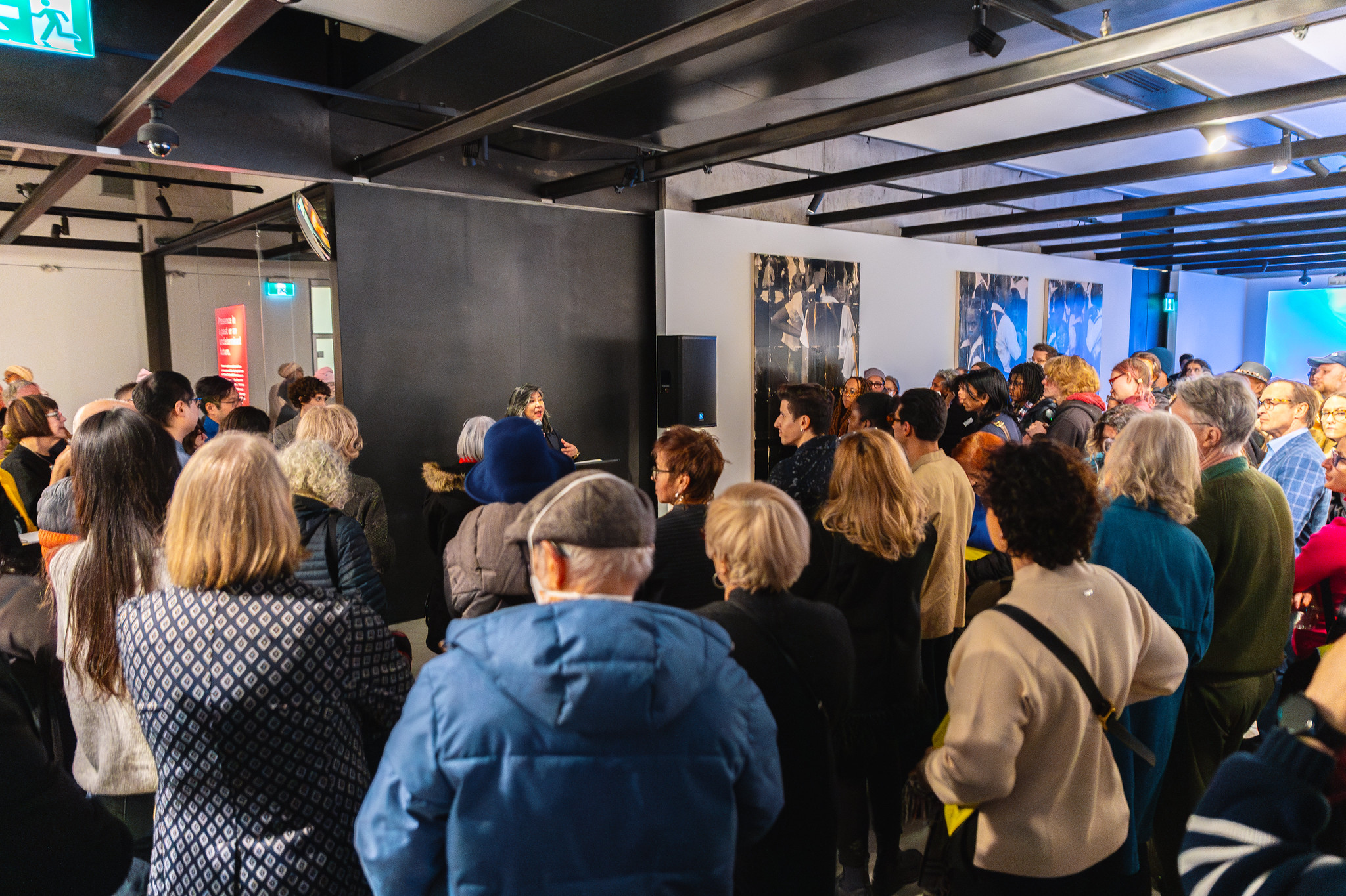 Large group of people in a gallery space listening to someone talk at a mic