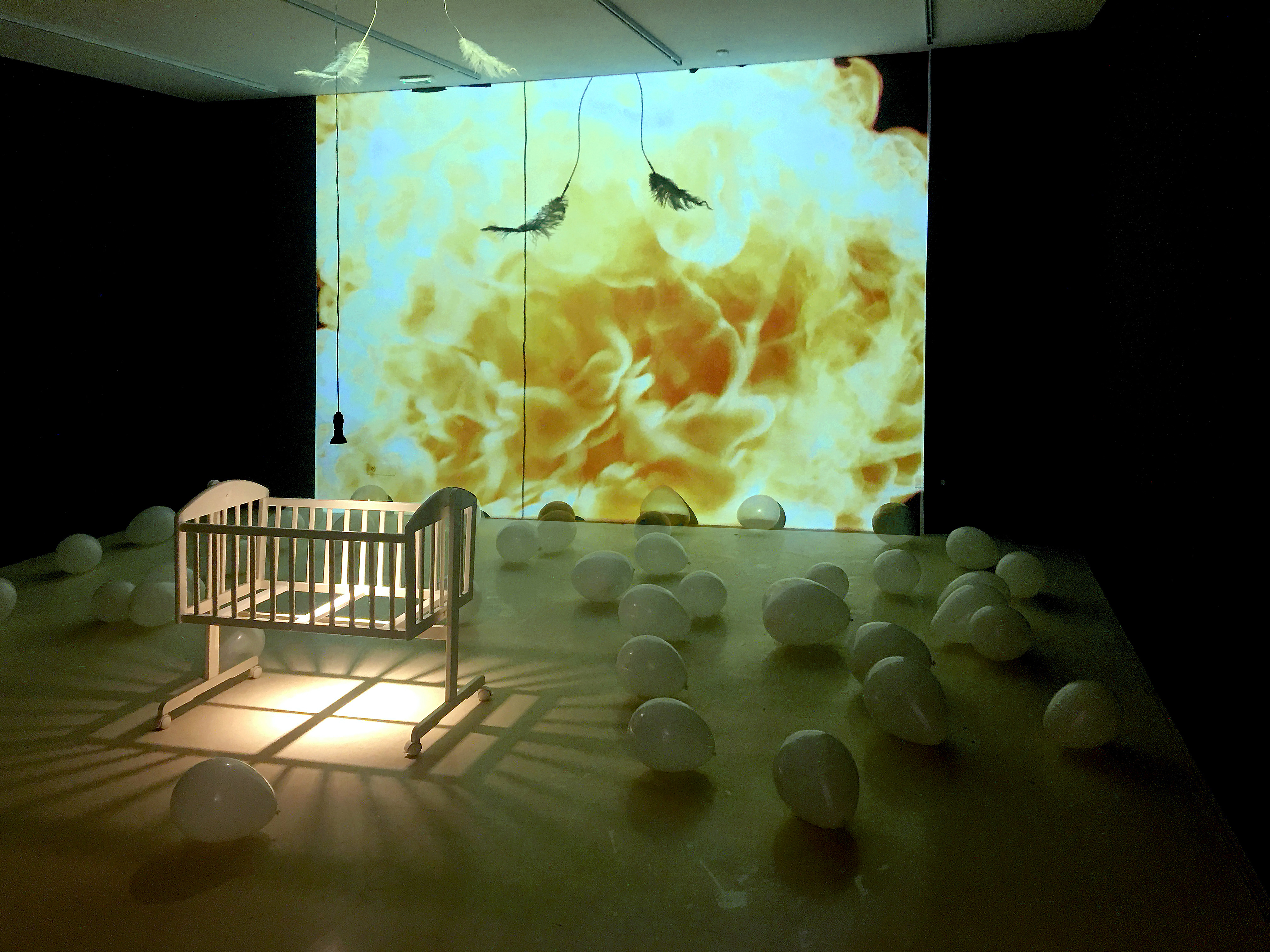 Video and sculptural installation, white wooden cradle, white ostrich feathers, balloons, 2 video projections: projection on the wall 4:03 video loop, sound; projection on the TV monitor 0:49 video loop, sound, dimensions variable.