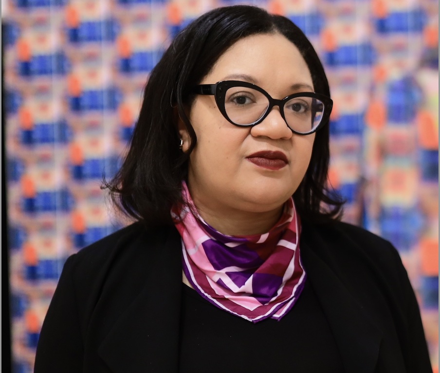 Alt. text: A woman with shoulder-length wavy black hair wearing black-framed eyeglasses, a purple striped neck scarf and a black shirt.