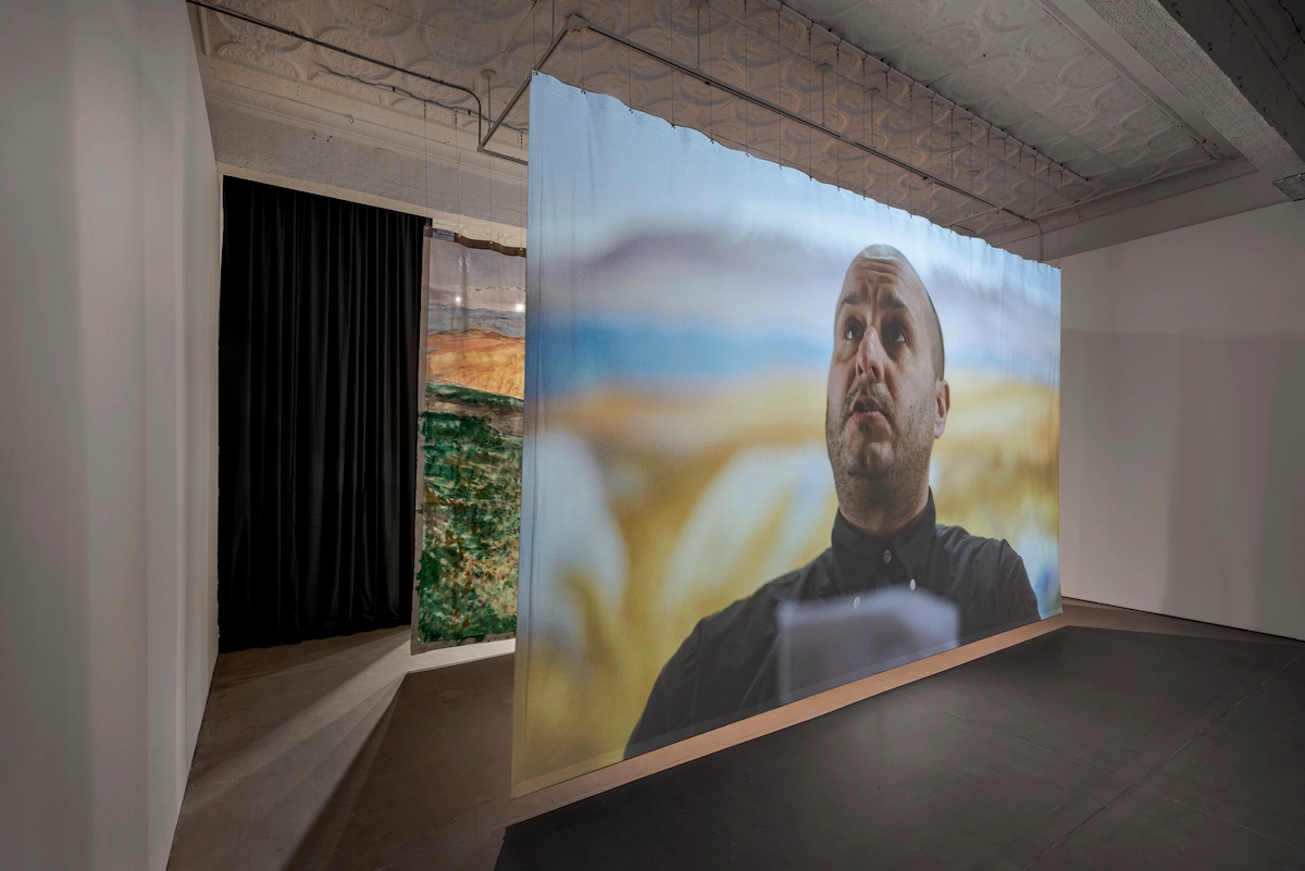 Photo of a room with two screens with projected images, one behind the other. The frontmost screen depicts a man looking up, with a landscape behind him.