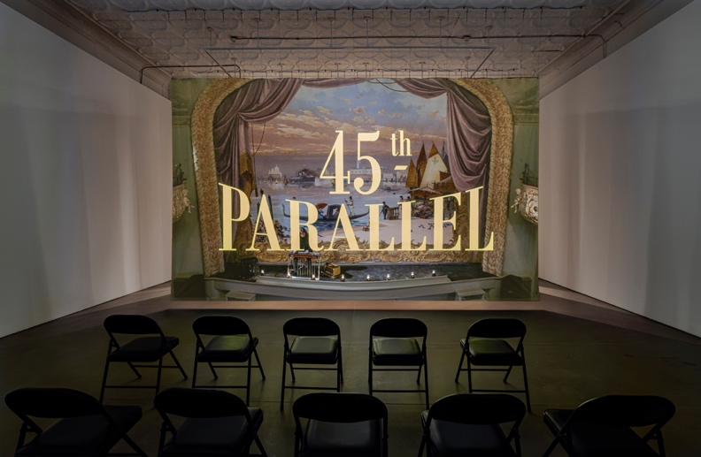 Photograph of a large projected image that reads "45th PARALLEL". Black metal chairs face the screen.
