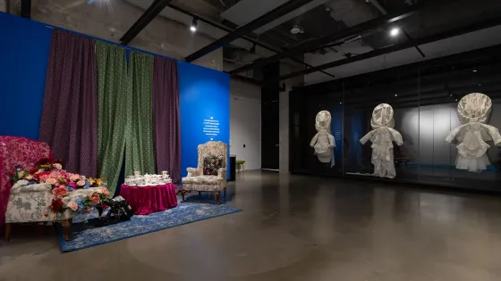 Installation view of Taqralik Partridge: ᐳᓛᖃᑎᒌᑦ (Pulaaqatigiit), Two floral chairs, one chair has artificial flowers, the other has a cushion, inviting visitors to sit. A small table has tea cups stacked on top, in front of floral curtains and a blue wall. On the right, Taqralik Partridge, qangattasuut (they fly).