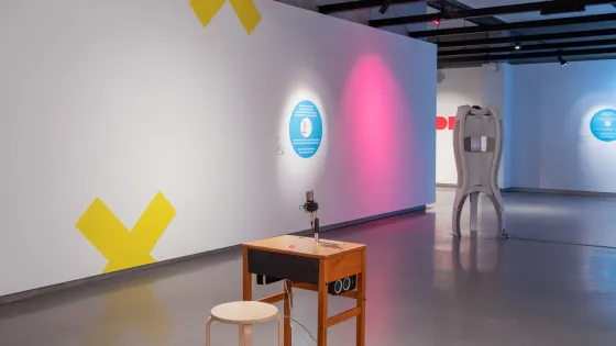 Installation view: CodeX: playable & disruptive futurist eArt, Onsite Gallery, OCAD University, Toronto, 2020. Photo: Yuula Benivolski. Artwork installation including yellow, blue and pink wall elements, wooden desk and stool, white structure. 