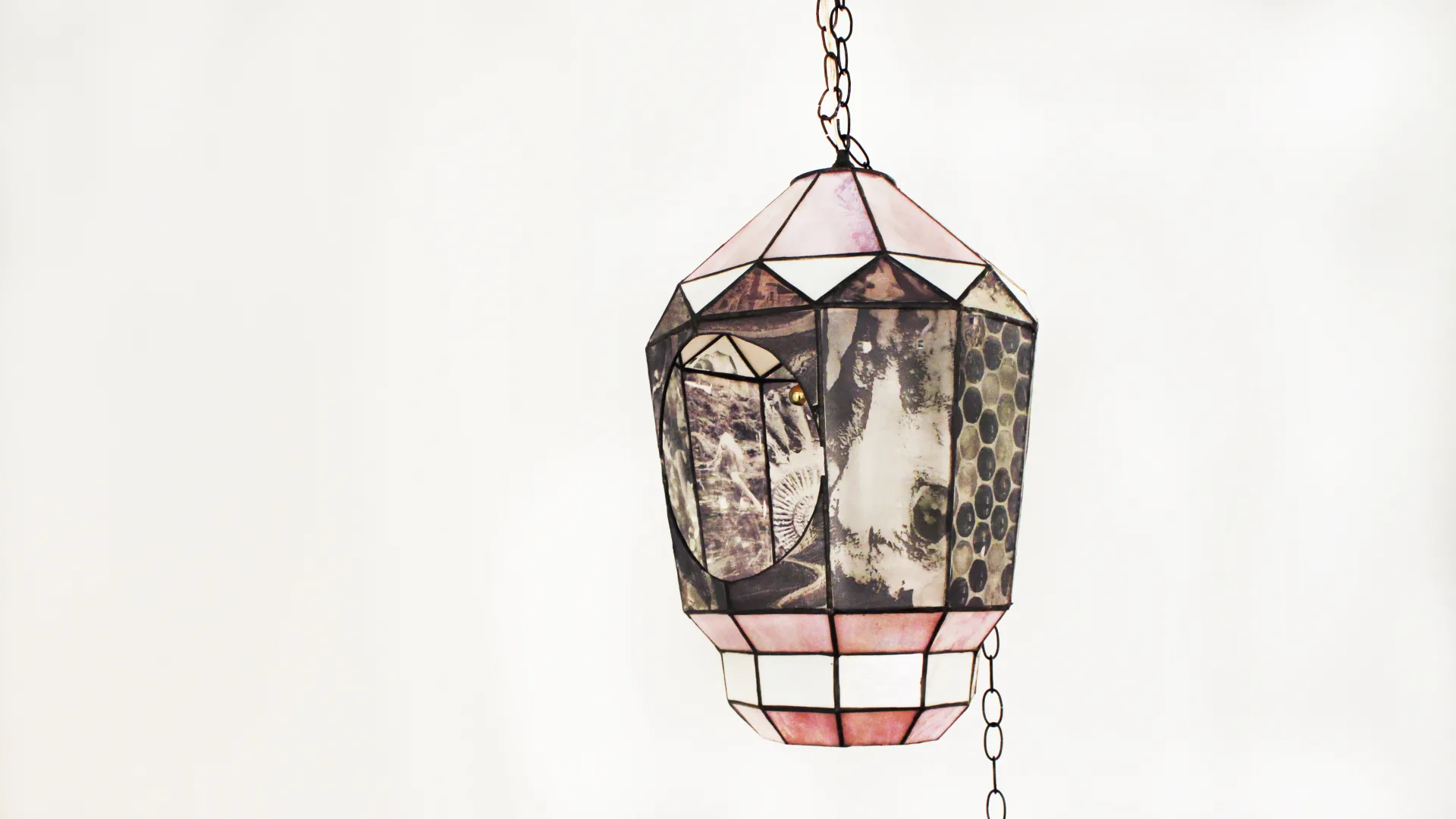 A stained glass lantern suspends from a long chain in front of a white wall. The lantern is covered in panels with black and white image transferred on the glass. There is a large circular opening to peer inside the lantern. 