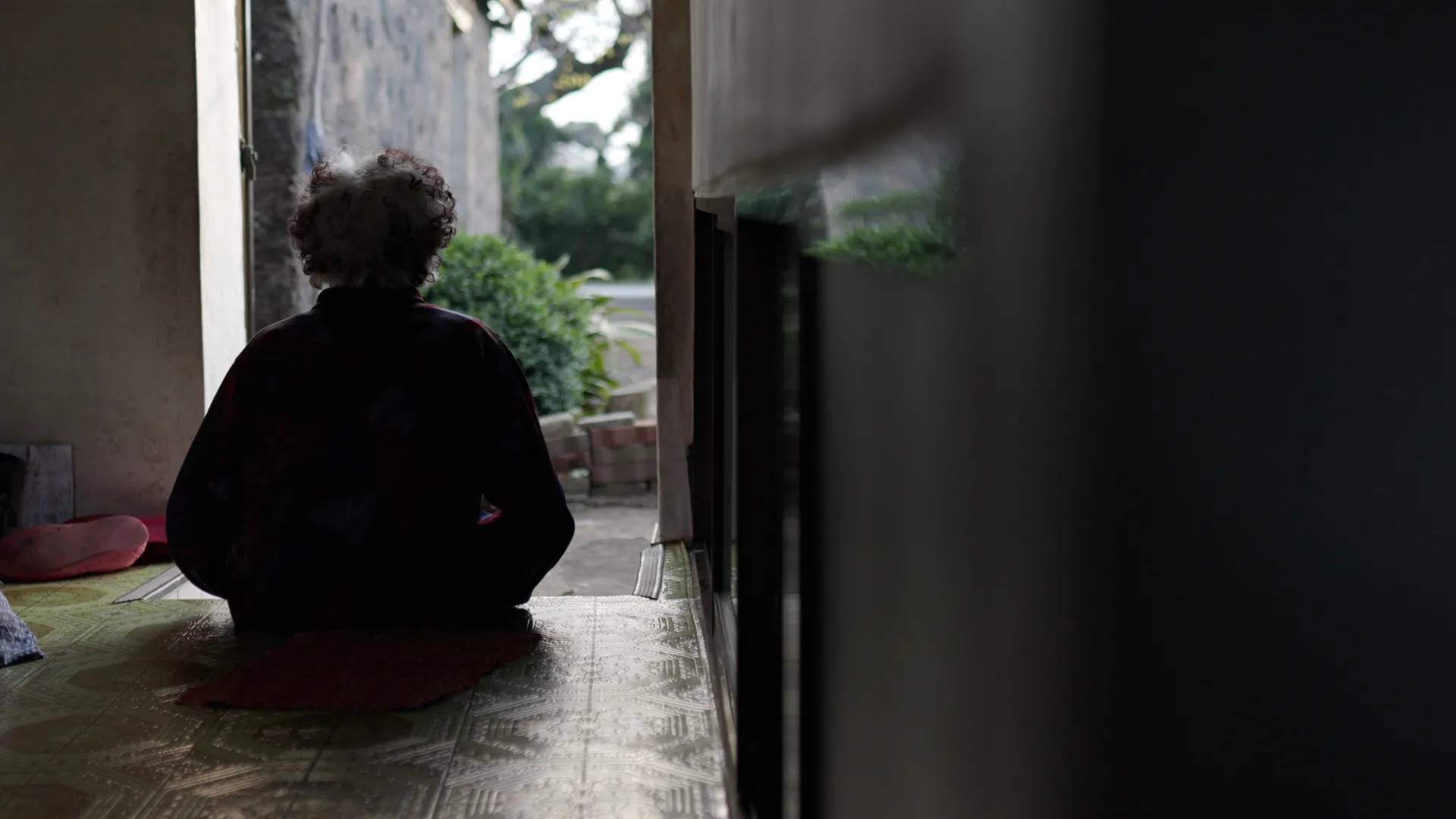 Film still of an Asian senior female sitting on the floor with backlighting