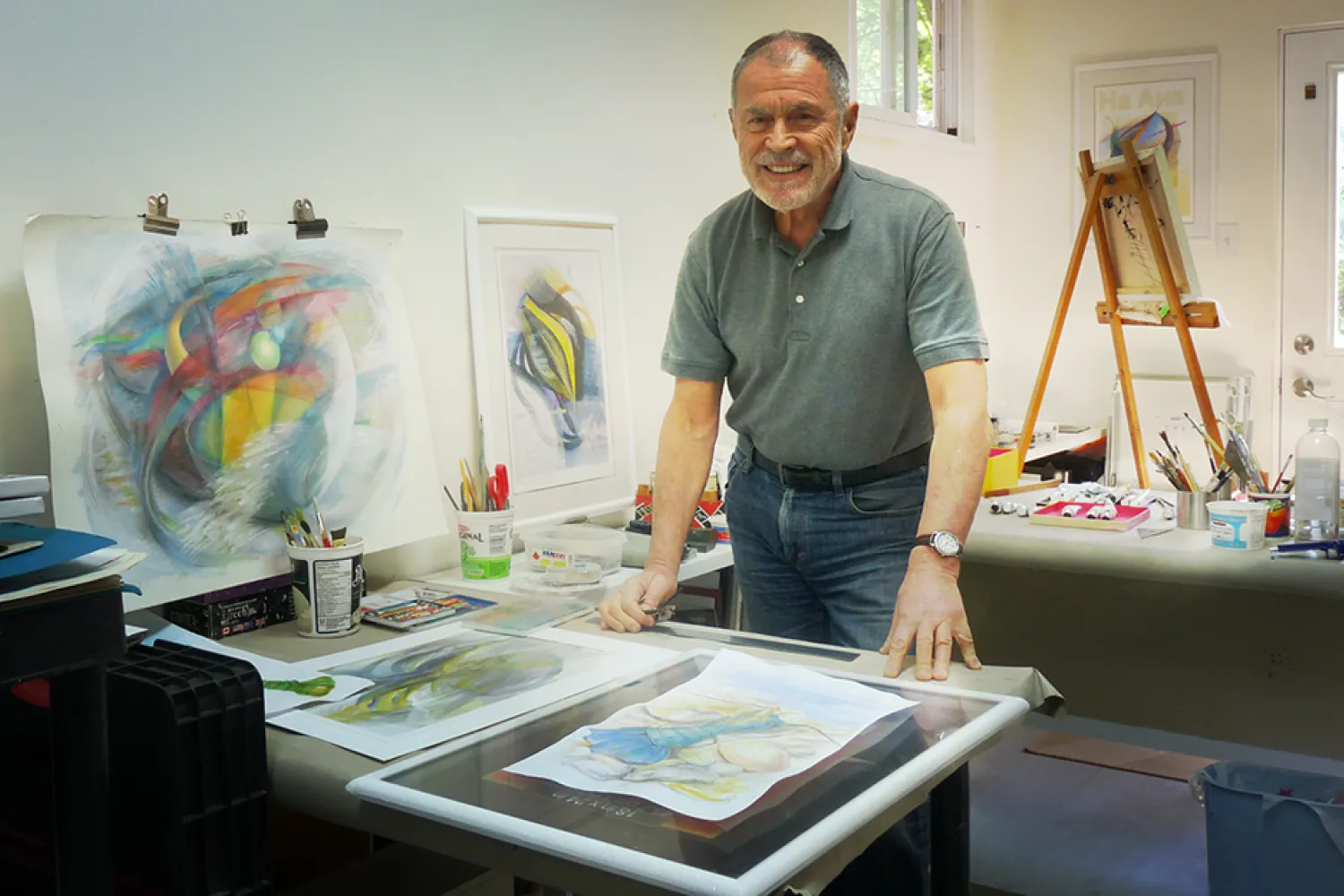 A photo of a smiling man wearing a greent shirt and blue jeans. He is standing in front of artprints and beside an easel in an art studio.