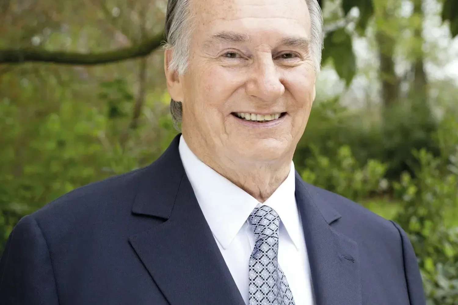 Photo of The Aga Khan wearing a navy suit , white shirt and blue and white patterned tie. 