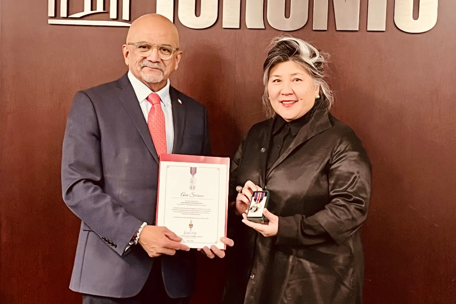 At left, man in a grey suit and red tie holding a certificate with the the city of Toronto in the background and the words, Toronto, in the background; at right, a woman with grey and black hair wearing a black jacket and pants holding a medal