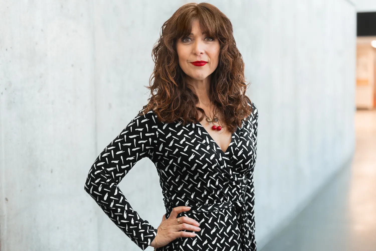 Woman with long brown hair wearing a black and white patterned dress stands with her hand on her. hip