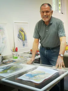 A photo of a smiling man wearing a greent shirt and blue jeans. He is standing in front of artprints and beside an easel in an art studio.