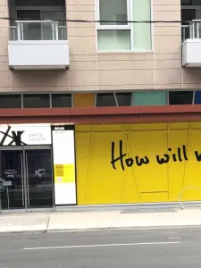 How will we be with you written in black on yellow background painted on window of the onsite gallery