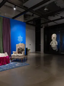 Installation view of Taqralik Partridge: ᐳᓛᖃᑎᒌᑦ (Pulaaqatigiit), Two floral chairs, one chair has artificial flowers, the other has a cushion, inviting visitors to sit. A small table has tea cups stacked on top, in front of floral curtains and a blue wall. On the right, Taqralik Partridge, qangattasuut (they fly).