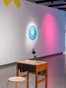 Installation view: CodeX: playable & disruptive futurist eArt, Onsite Gallery, OCAD University, Toronto, 2020. Photo: Yuula Benivolski. Artwork installation including yellow, blue and pink wall elements, wooden desk and stool, white structure. 