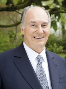 Photo of The Aga Khan wearing a navy suit , white shirt and blue and white patterned tie. 