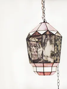 A stained glass lantern suspends from a long chain in front of a white wall. The lantern is covered in panels with black and white image transferred on the glass. There is a large circular opening to peer inside the lantern. 