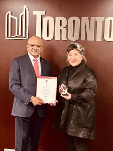 At left, man in a grey suit and red tie holding a certificate with the the city of Toronto in the background and the words, Toronto, in the background; at right, a woman with grey and black hair wearing a black jacket and pants holding a medal