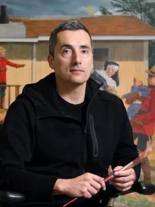 Colour photo of Kent Monkman holding a paintbrush, seated in front of his art work. 