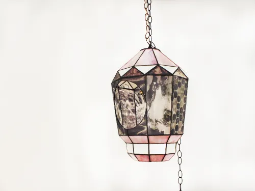 A stained glass lantern suspends from a long chain in front of a white wall. The lantern is covered in panels with black and white image transferred on the glass. There is a large circular opening to peer inside the lantern. 