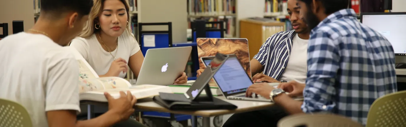 Four people sitting at a table with open laptobs in front of library stacks.