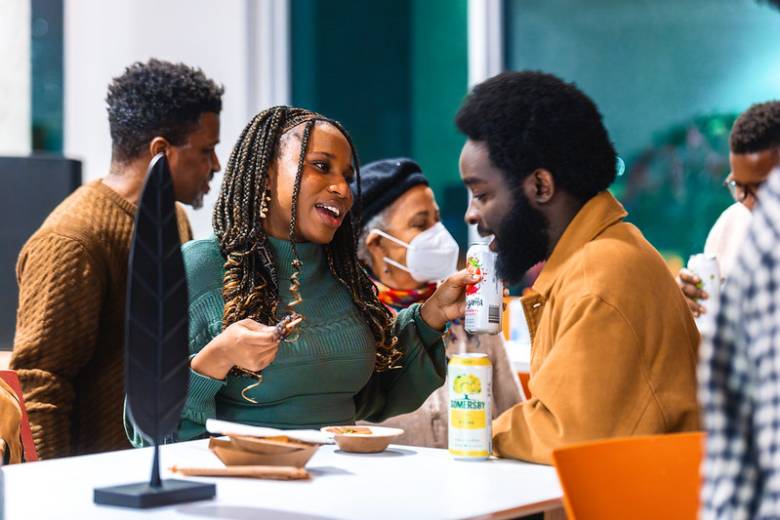 A photo of three Black people, a man with a beard at the left, a woman with long hair in the middle and a man with a beard at right wearing an orange shirt - they are at a high top table.