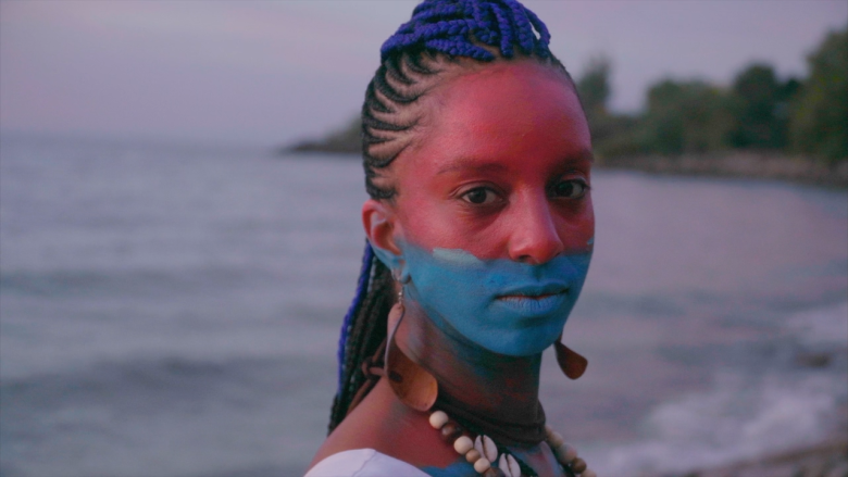 A black woman faces the camera with a painted face and a lake in the background.