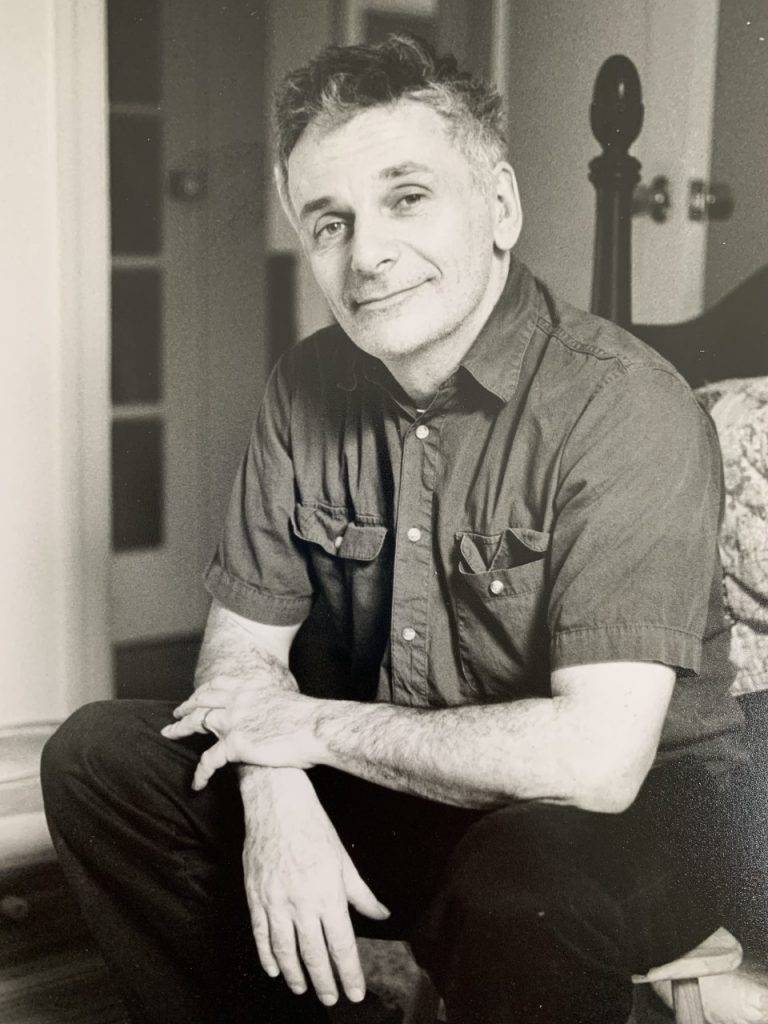 A black and white photo of a middle-aged man sitting in a living room smiling.