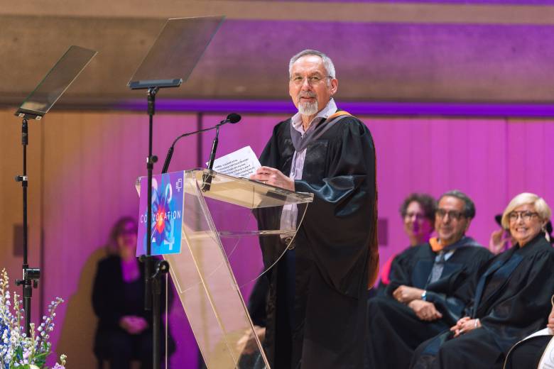 Photo of man with glasses at podium with grey hair, wearing a robe after receiving an honorary doctorate degree. Background has three people on stage watching him with a pinkish background.