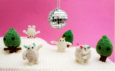 Pink background with a disco silver ball in the middle, there are 5 knitted off-white creatures with black eyes and red or green or yellow ears. One is standing on its head, two are waving and two are hugging. There are two knitted green trees and they are on an-off white knitted blanket.