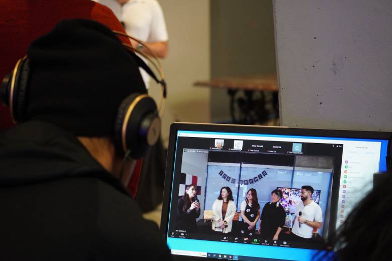 A photo of a man wearing headphones looking at a computer screen with five people, including from left to right four women and one man holding a microphone.